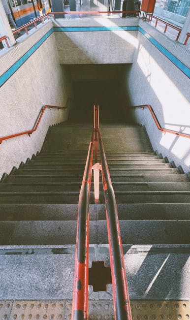 A downward view of a concrete staircase inside a building, with metal handrails painted in reddish-brown along both sides. The staircase leads to an underground or lower level, with ambient lighting illuminating the steps. The surrounding walls are finished in light grey with a textured finish, and there are small sections of wall with darker paneling. The scene is part of a building that could serve as a station or public space, and the image relates to the initial stages of home relocation or furniture transport, such as preparing to load items into a moving vehicle or navigating stairs during a move, in the context of professional removals like those offered by Man with Van Bromley.
