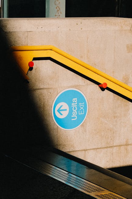 A blue circular exit sign with the word 'Uscita' and an upward arrow is mounted on a beige stone wall at Bromley North Station. An escalator with a yellow handrail and black safety stripe descends alongside the wall, with red safety stops securing the railing. Below the sign, a platform area features a metal edge and a textured strip for safety. This scene illustrates the station's interior, highlighting the functionality of the exit signage and the escalator used for home relocation and moving logistics at this busy transport hub, as part of the services offered by Man with Van Bromley.