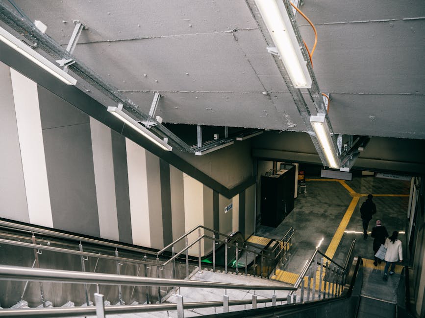 A downward view of a concrete staircase inside a building, with metal handrails painted in reddish-brown along both sides. The staircase leads to an underground or lower level, with ambient lighting illuminating the steps. The surrounding walls are finished in light grey with a textured finish, and there are small sections of wall with darker paneling. The scene is part of a building that could serve as a station or public space, and the image relates to the initial stages of home relocation or furniture transport, such as preparing to load items into a moving vehicle or navigating stairs during a move, in the context of professional removals like those offered by Man with Van Bromley.