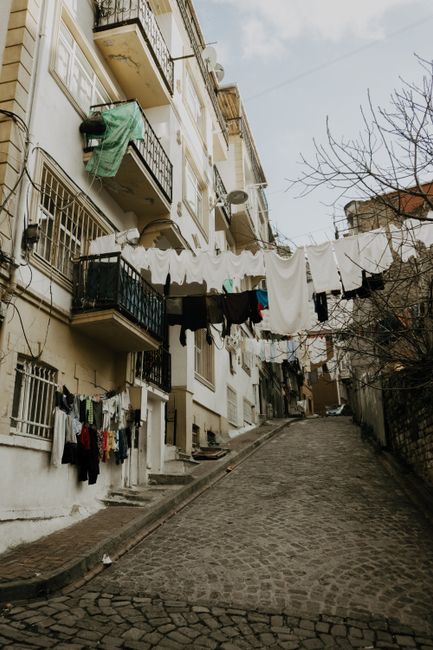 A narrow cobbled street lined with multi-storey residential buildings featuring small balconies with metal railings. Laundry including shirts, trousers, and towels are hung on clotheslines strung across the street, attached to the buildings' facades. Some clothing items are draped over the rails or clipped onto the lines. The street slopes upward and is illuminated by natural daylight, with bare tree branches visible on the right side. This scene depicts an everyday residential area, typical of Tightlands properties, where external clothes drying is common, and it erails the environment in which house relocation and moving services by Man with Van Bromley could be coordinated, especially in tight street conditions for furniture transport and home relocation logistics.